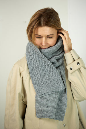 A woman wearing a wide hand-knitted scarf in Cool Grey over a cream coat.