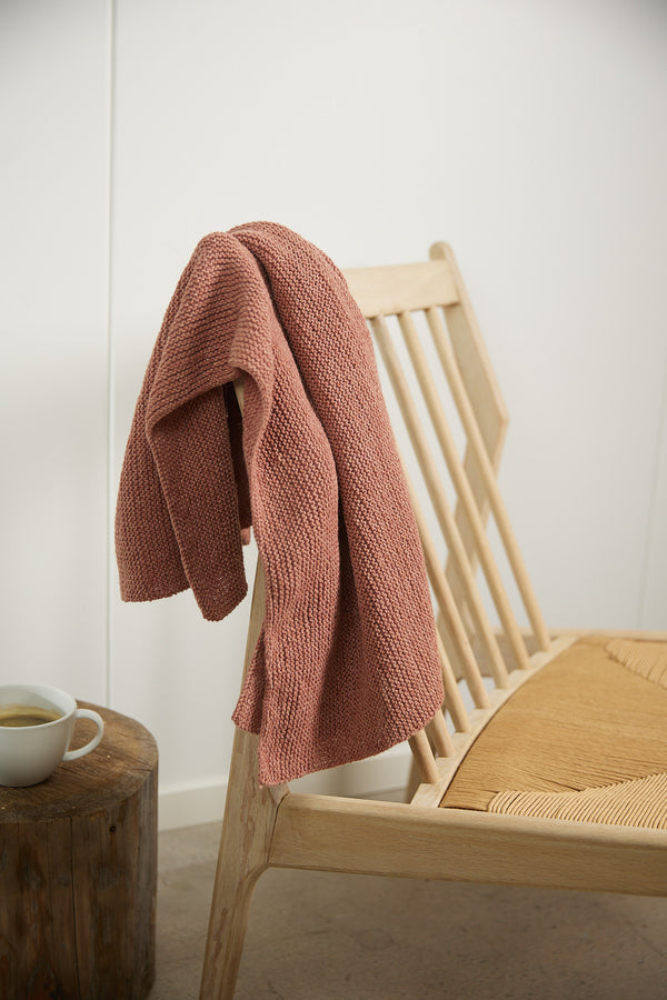 A finished scarf in Rose draped over a light wooden chair, next to a cup of coffee.