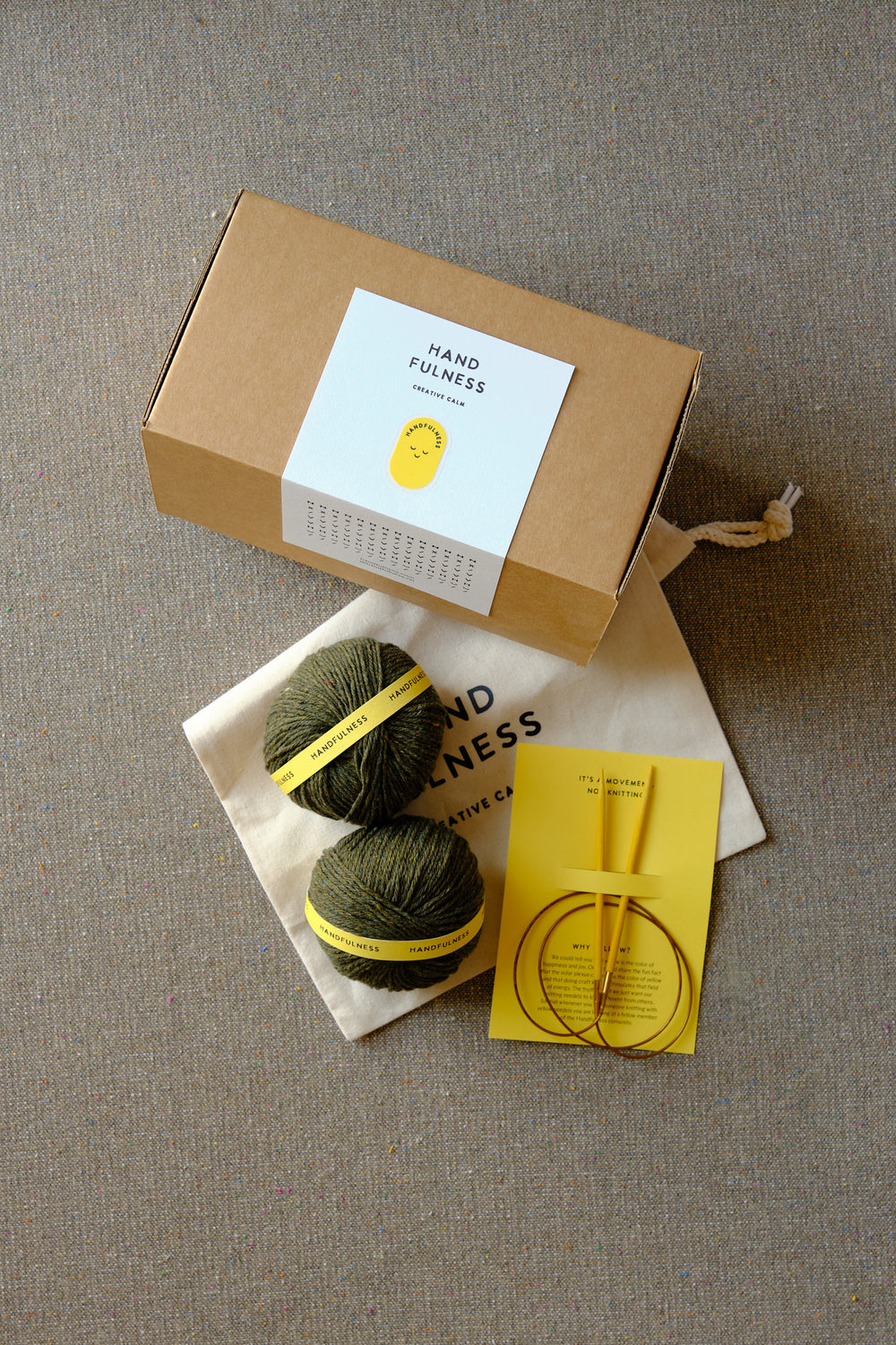 Small Handfulness kit in Forest Green: two balls of dark green yarn, wooden needles, a cotton bag and a kraft box.