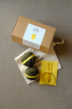 Small Handfulness kit in Forest Green: two balls of dark green yarn, wooden needles, a cotton bag and a kraft box.