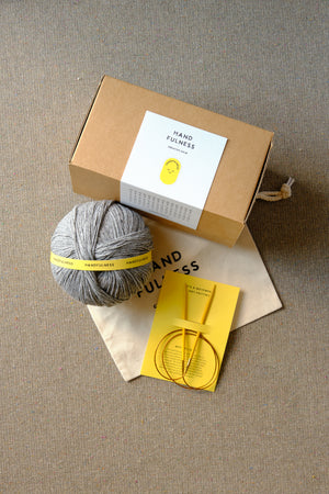 Medium Handfulness kit in Cool Grey: a ball of grey yarn, wooden needles, a cotton bag and a kraft box.