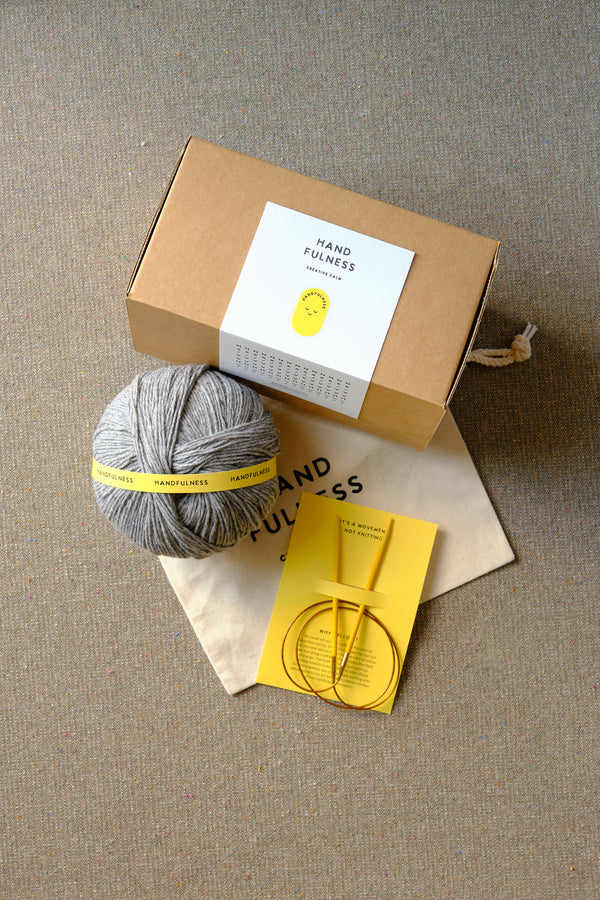 Medium Handfulness kit in Cool Grey: a ball of grey yarn, wooden needles, a cotton bag and a kraft box.