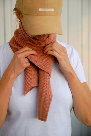 A woman tying a hand-knitted scarf in Rose over a white t-shirt.