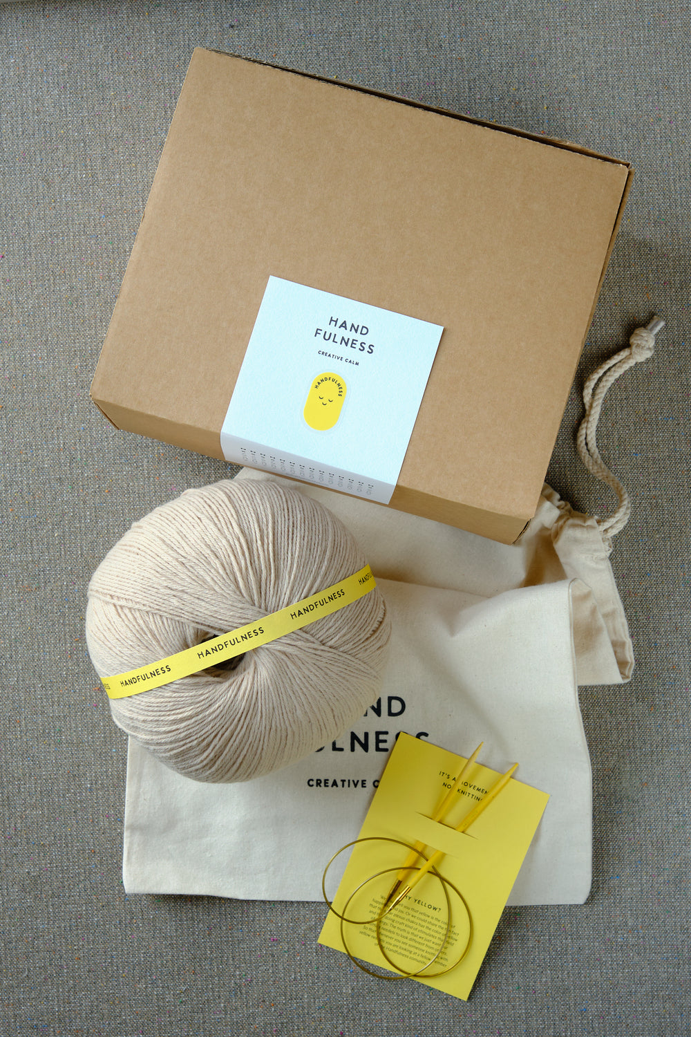 Large Handfulness kit in Sand: a ball of beige yarn, wooden needles, a cotton bag and a kraft box.