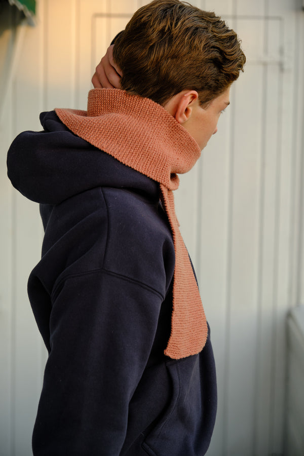 A man wearing a hand-knitted scarf in Rose over a dark hoodie.